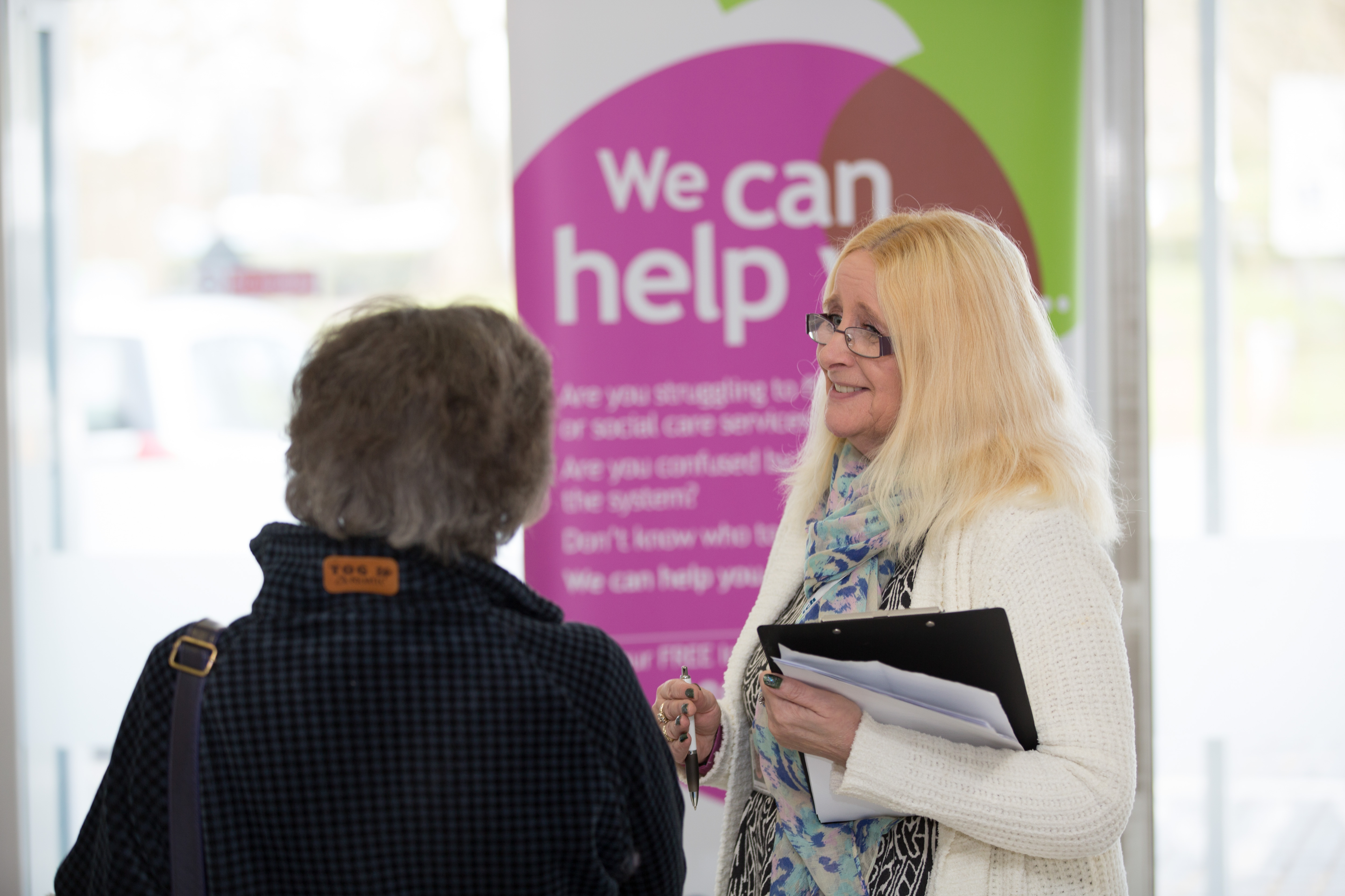 Woman standing in front of a banner that says 'we can help' speaking to a member of the public